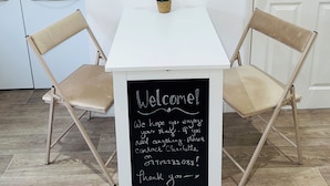 A small, white table with a chalkboard sign reading "Welcome! We hope you enjoy your stay. If you need anything, please contact Charlotte on 07912282088. Thank you." Two fold-up chairs are positioned on either side of the table. Above the table, there is a shelf with two candles and a small plant. - Wanstead Gem – Bright, Cosy & Immaculate High Street Apartment in East London (London)