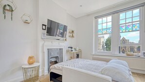 A modern bedroom with a bed, a TV, a fireplace, and a large window with a view of a building. - Wanstead Gem – Bright, Cosy & Immaculate High Street Apartment in East London (London)