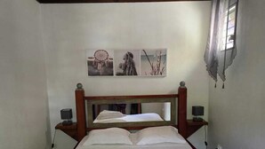 A wooden bed with white linens and a headboard, a bedside table with a lamp, and a wall with three framed pictures. - Bungalow Le Refuge Vert (Morne-à-l'Eau)