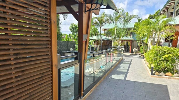 A covered walkway with a glass balustrade, leading to a pool area with a view of a tropical resort. - Bungalow Le Refuge Vert (Morne-à-l'Eau)