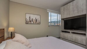 A hotel room with a bed, a TV, a painting, and a window with a city view.