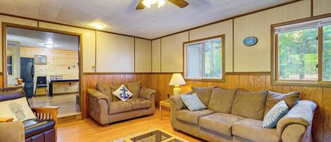 A cozy living room with a ceiling fan, wooden walls, and a large window.
