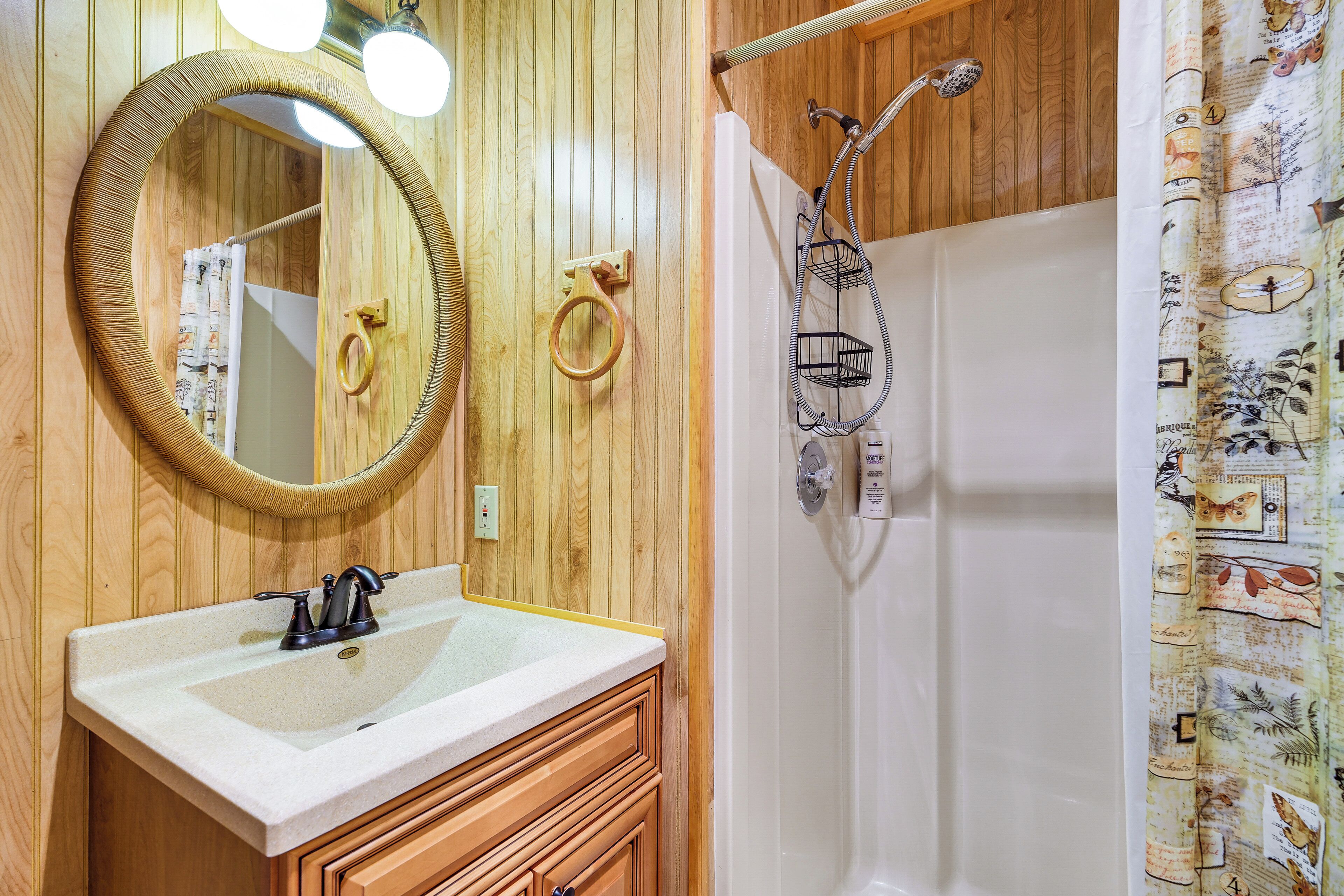 A bathroom with a wooden vanity, a round mirror, a shower with a hose, and a curtain with a vintage pattern.