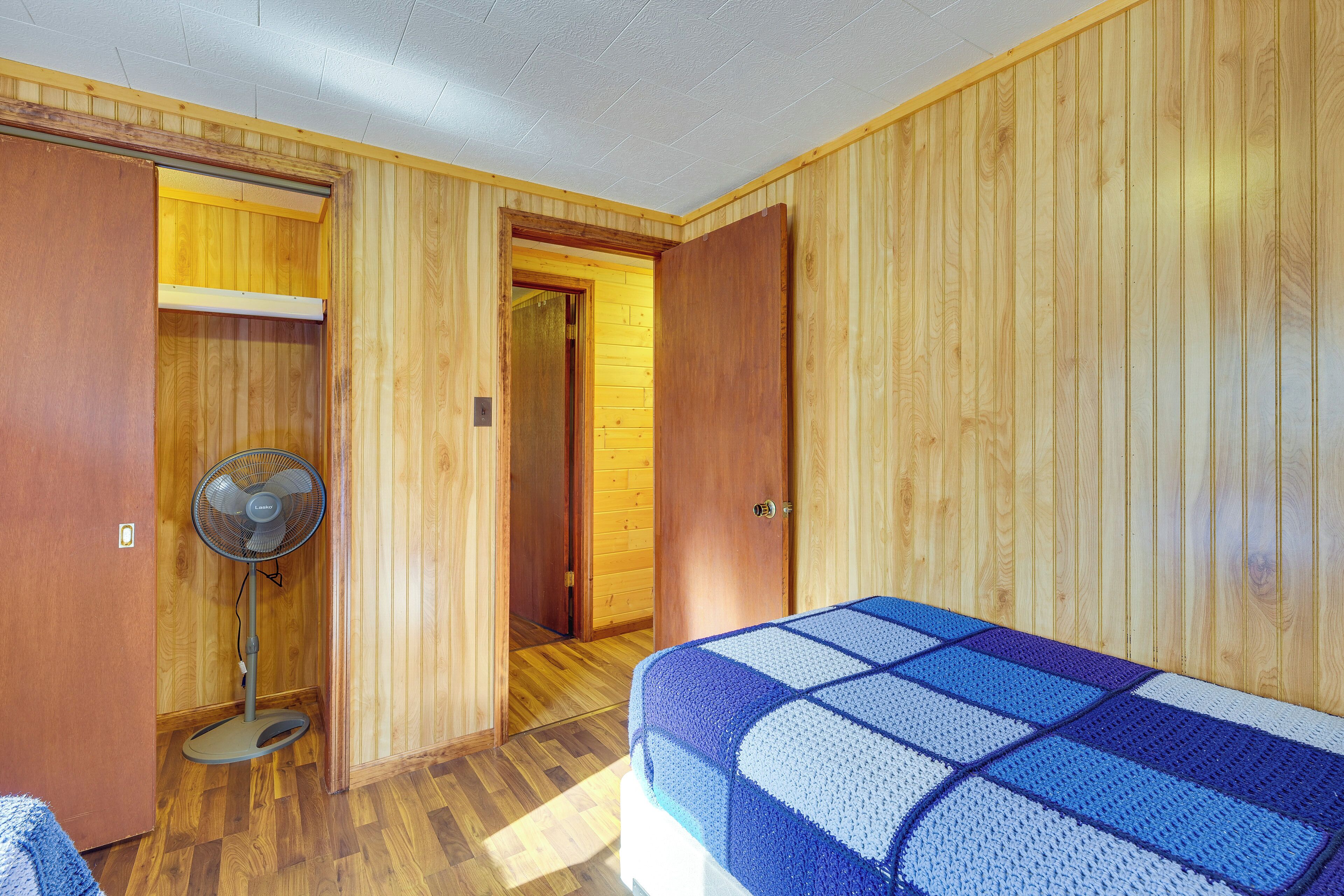 Une pièce aux murs recouverts de lambris en bois, un lit avec une couverture bleue et blanche, un ventilateur sur pied et une porte donnant sur une autre pièce.