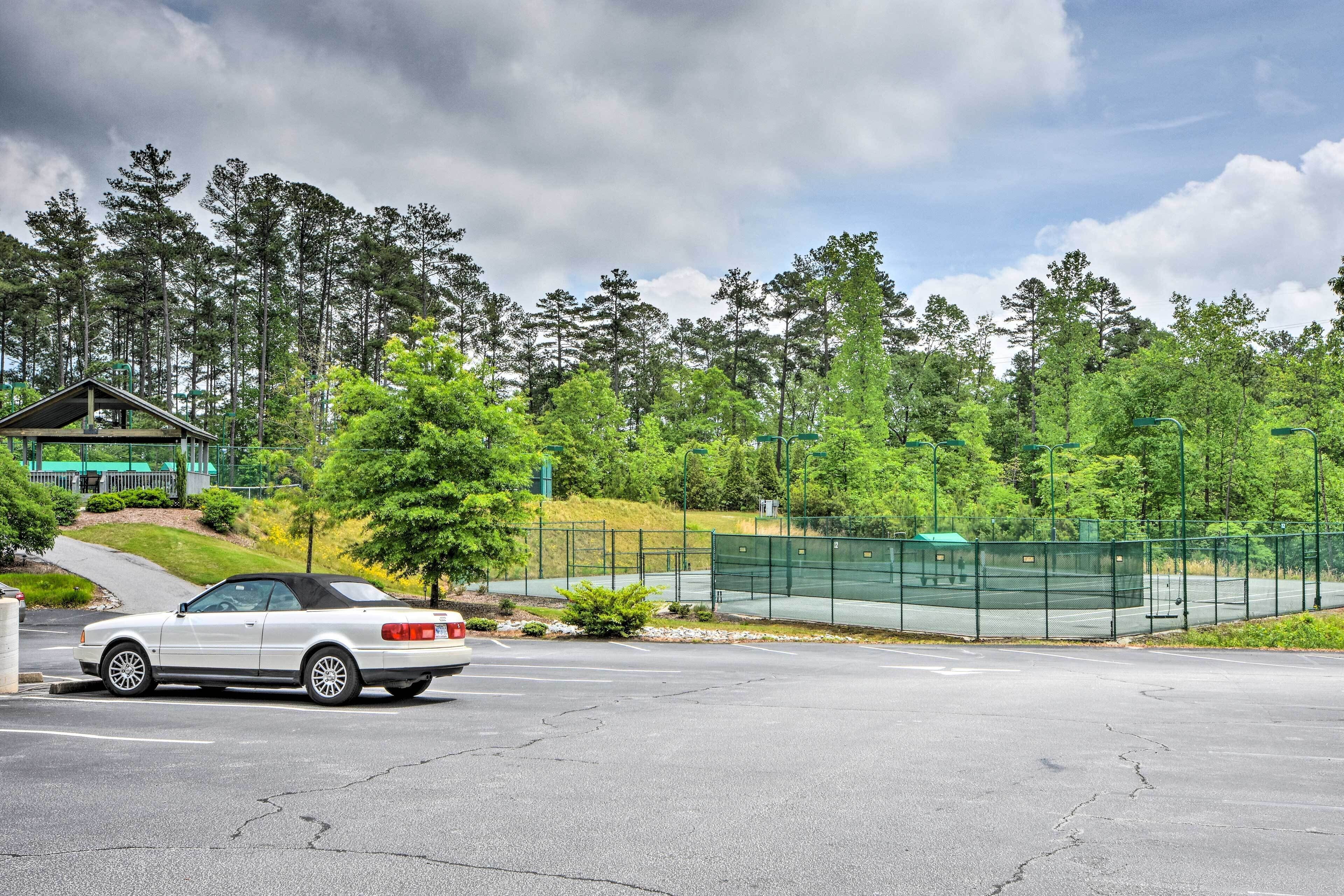 A white convertible car parked in a parking lot with a tennis court and trees in the background.