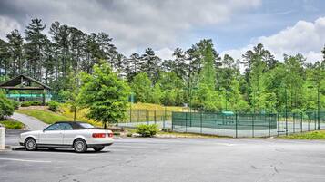 A white convertible car parked in a parking lot with a tennis court and trees in the background.