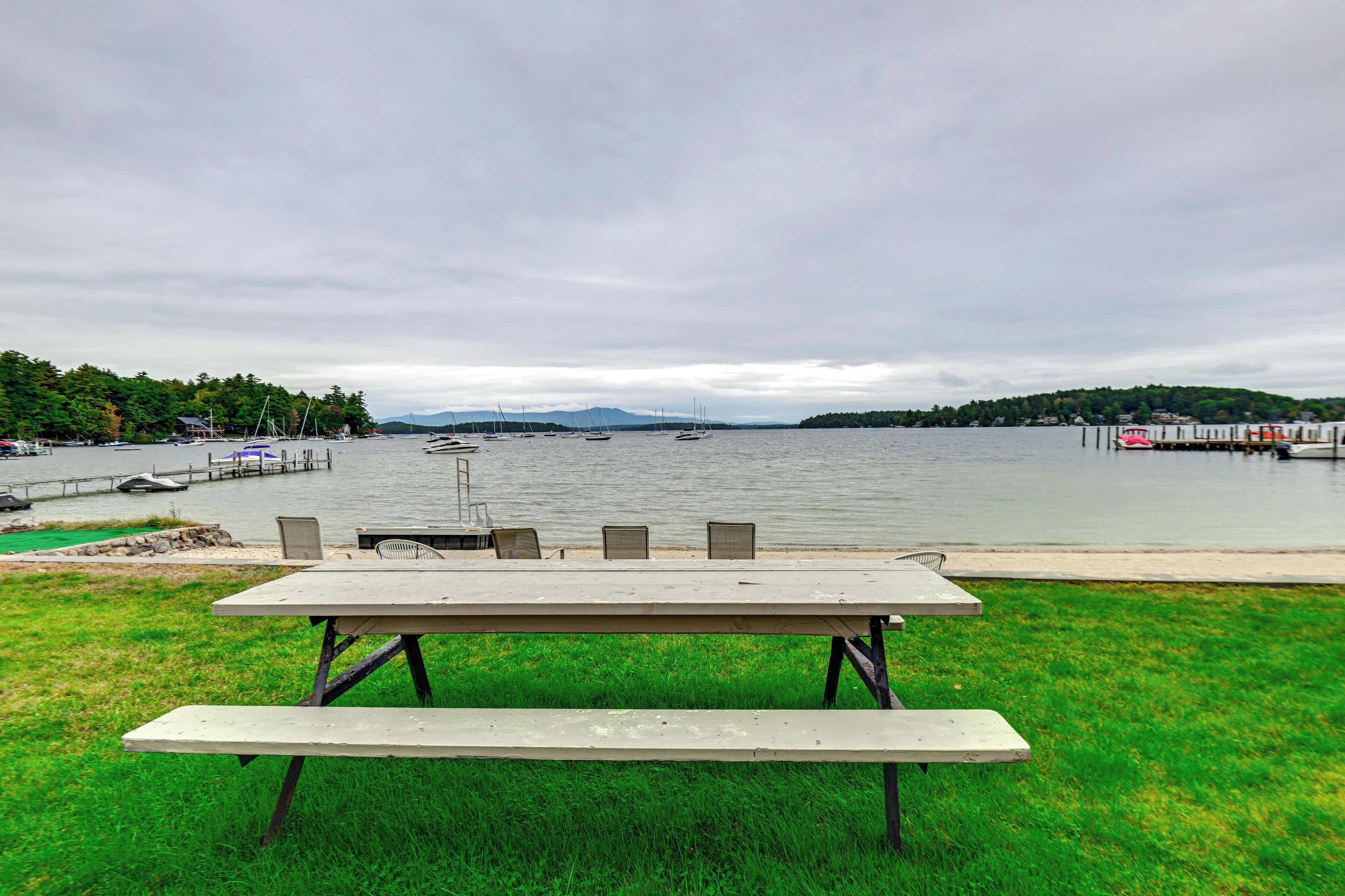 A picnic table with chairs in a grassy area overlooking a lake with boats and a dock.