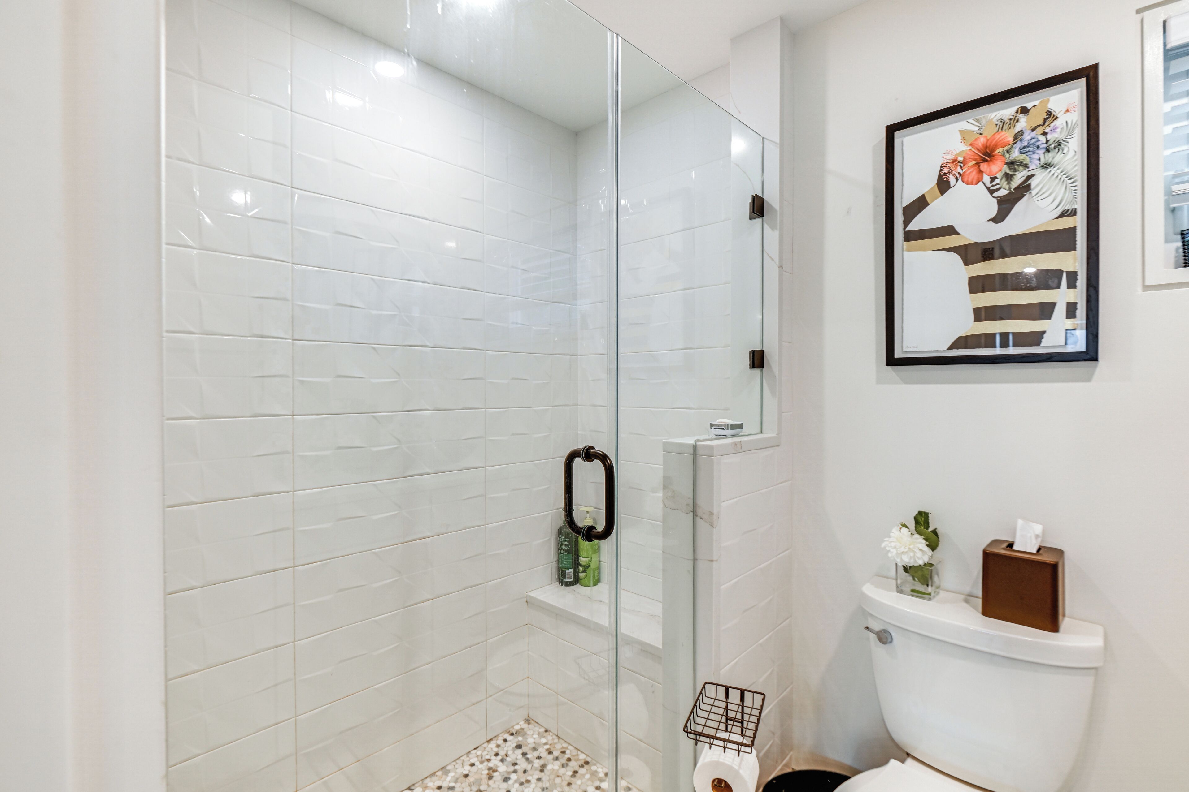 A modern bathroom with white tiled walls, a glass shower enclosure, a toilet with a tissue box and a small vase of flowers, and a framed artwork above the toilet.