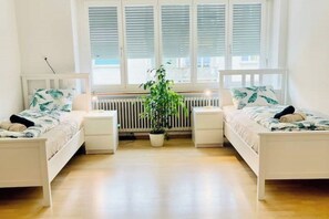 Ein Zimmer mit zwei Betten, einem Heizkörper, einem Topf mit Pflanze und einem Fenster mit Jalousien.