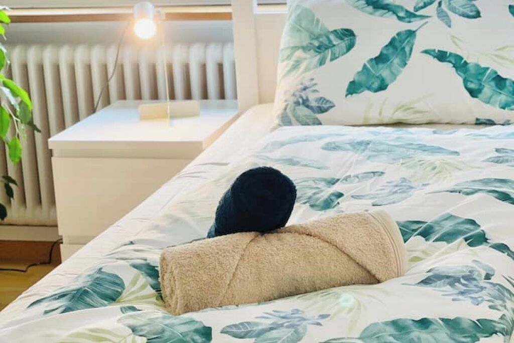 Een opgemaakt bed met een tropisch bladmotief, een nachtkastje met een lamp, en een radiator op de achtergrond.