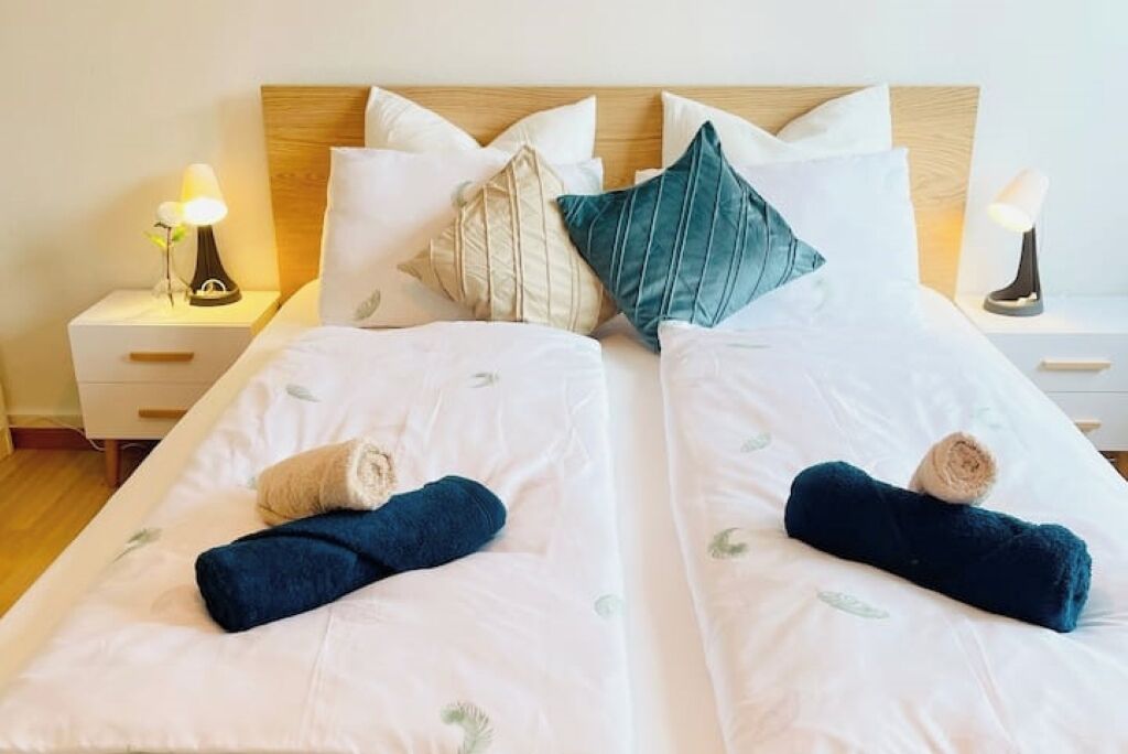 A double bed with white bedding and two pillows, a nightstand with a lamp, and a wooden headboard.