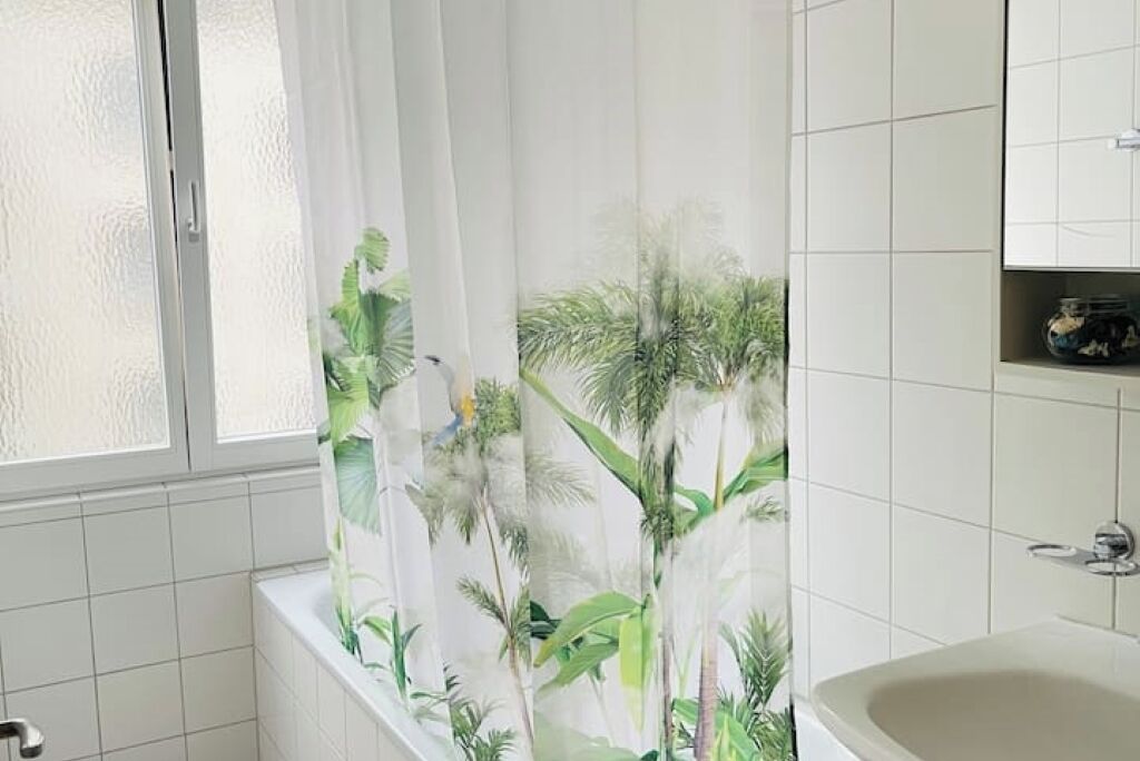 A bathroom with a bathtub, a window with frosted glass, and a shower curtain with a tropical leaf pattern.
