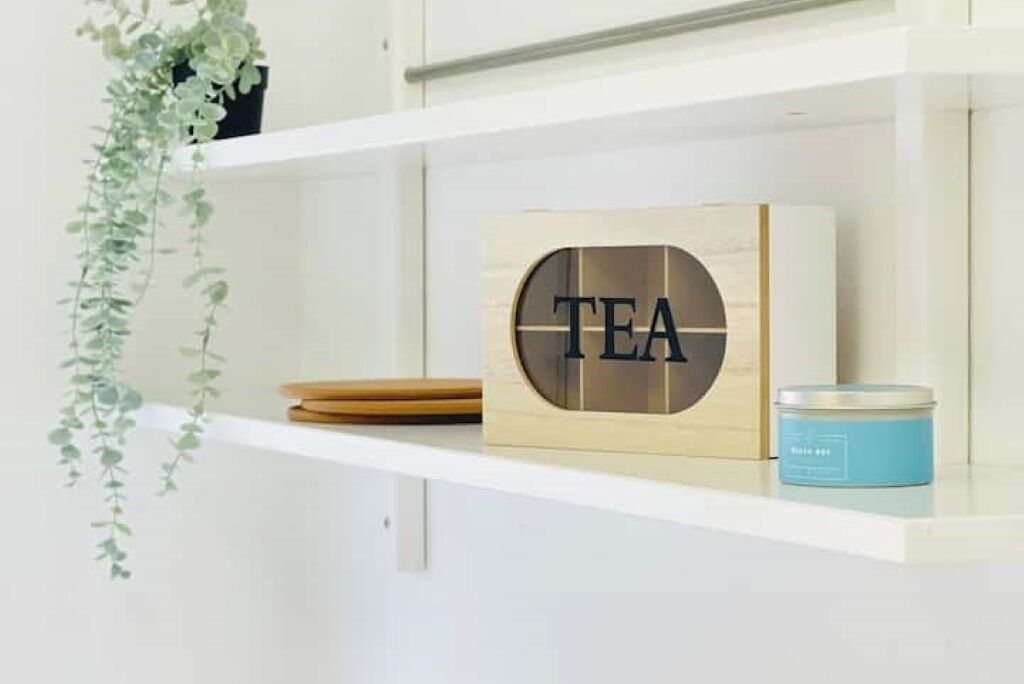 A shelf with a wooden block displaying the word "TEA," a small blue container, and a hanging plant.