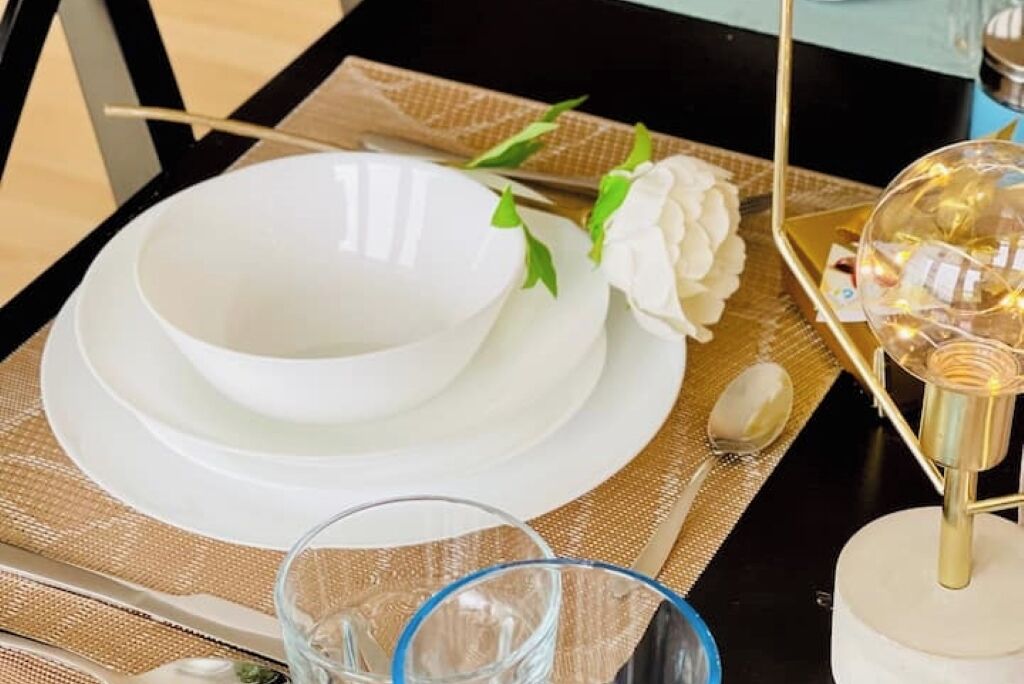 A table set with white dishes, a clear glass, and a gold tray with a light fixture.