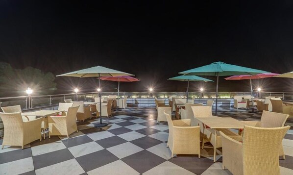An outdoor patio with checkered flooring, wicker furniture, and colorful umbrellas. - Hotel Woodville Inn - Hotel in Mcleodganj  (Dharamshala)