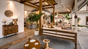 A spacious living area with a wooden bench, a round wooden table, and a white sofa. There is a large potted plant and a wicker chair in the background. - Villa Folie by Alfred in Bali - New 5bdr Dream Villa in the Heart of Seminyak (Seminyak)