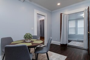 A dining area with a round table, grey chairs, a potted plant, and a view of a window with curtains. - Fully Stocked Kitchen,  AC,  Wi-Fi, W/D,  Near Baseball and Walk to Downtown (Oneonta)