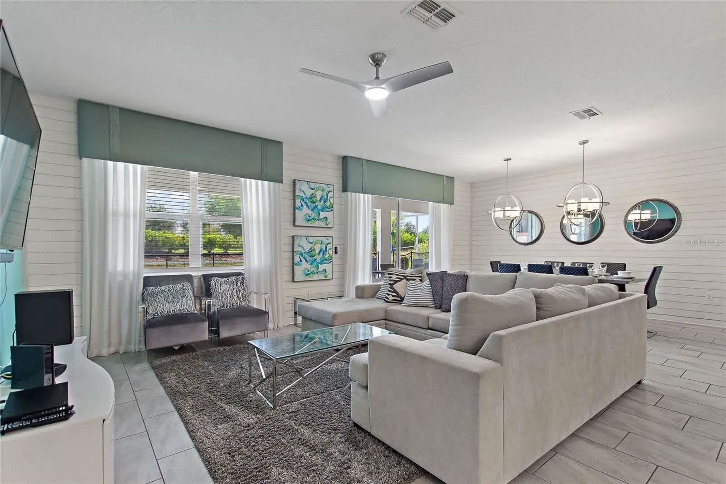 A spacious living room with a large sofa, a glass coffee table, and a flat-screen TV. There are two windows with curtains, a ceiling fan, and artwork on the walls.