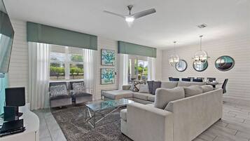 A spacious living room with a large sofa, a glass coffee table, and a flat-screen TV. There are two windows with curtains, a ceiling fan, and artwork on the walls.