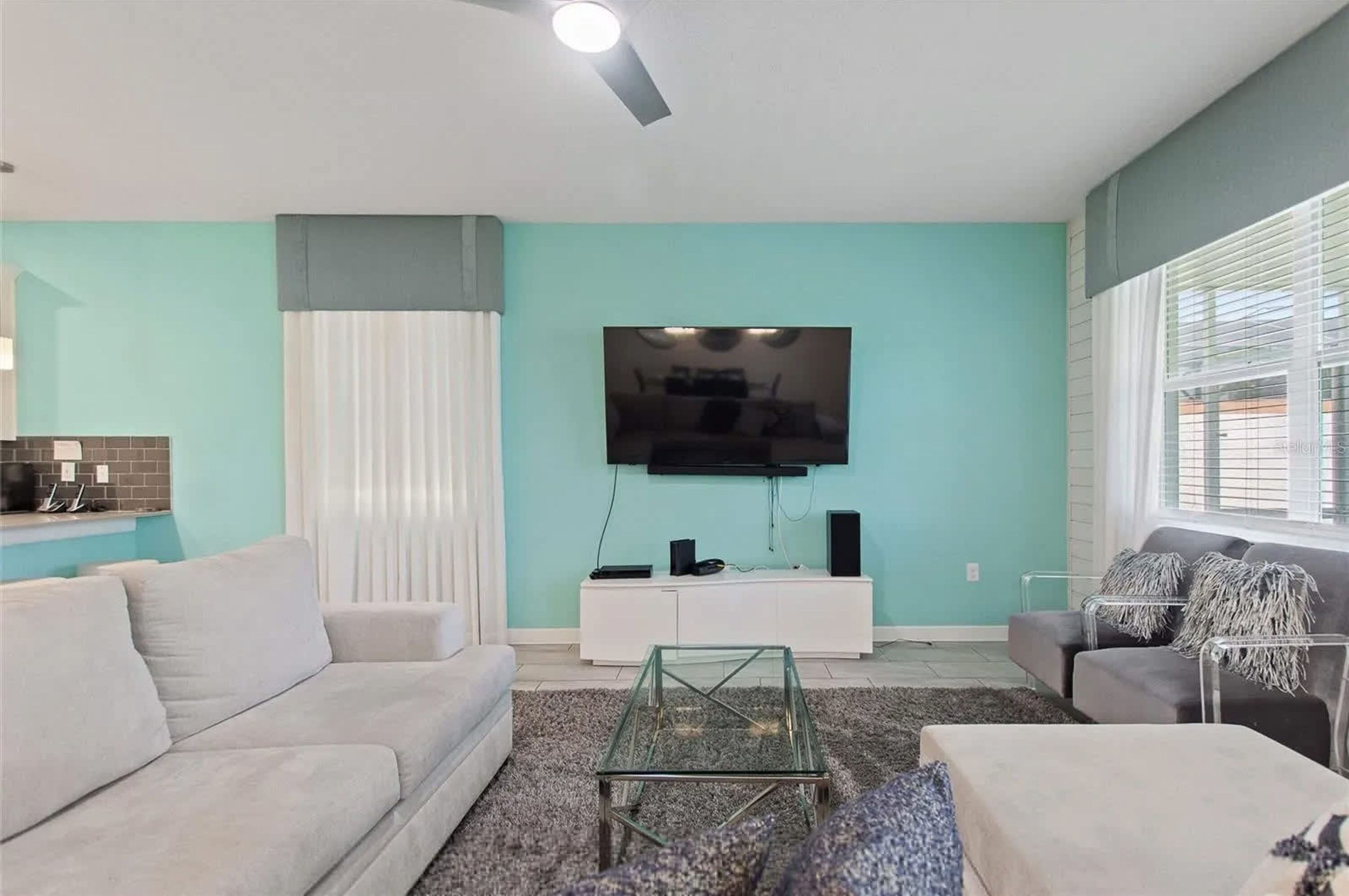 Uma sala de estar com parede verde-água, uma TV de tela plana montada acima de um móvel branco, um sofá cinza, uma mesa de centro de vidro e uma janela com persianas.