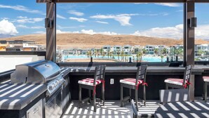 A rooftop bar with a grill, chairs, and a view of a pool and buildings.