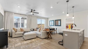 A modern living room with a sofa, ottoman, and coffee table, a dining area with a table and chairs, a kitchen with a sink and countertop, and a ceiling fan.
