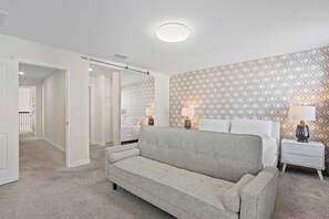 A bedroom with a grey sofa, a white bedside table with lamps, and patterned wallpaper.