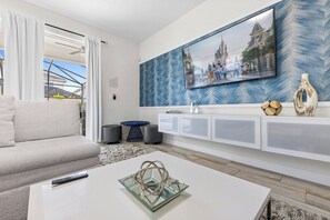 A modern living room with a sofa, a coffee table, a TV mounted on a wall with a decorative painting, and a white console unit with storage.