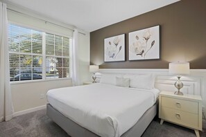 A hotel room with a large bed, two bedside tables, a window with blinds, and two wall-mounted paintings. - Kissimmee Bliss: 6BR Pool Home w Game Room (Kissimmee)