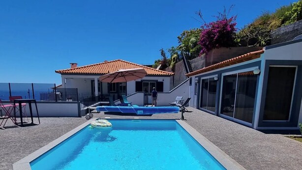A rectangular pool with clear blue water, surrounded by a stone deck. There are lounge chairs and a small inflatable float. The pool is adjacent to a building with a tiled roof and large windows. - Casa Agapanthe House  (Estreito da Calheta)