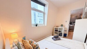 A bedroom with a bed, a bedside lamp, a window, a shelf with books and a plant, and a view of the kitchen.