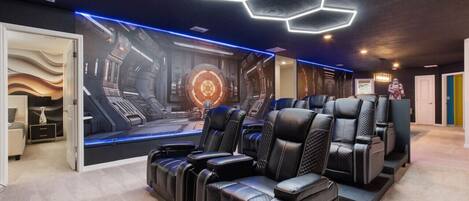 A room with a futuristic design, featuring a large mural of a space station, black leather chairs, and a hexagonal light fixture on the ceiling.