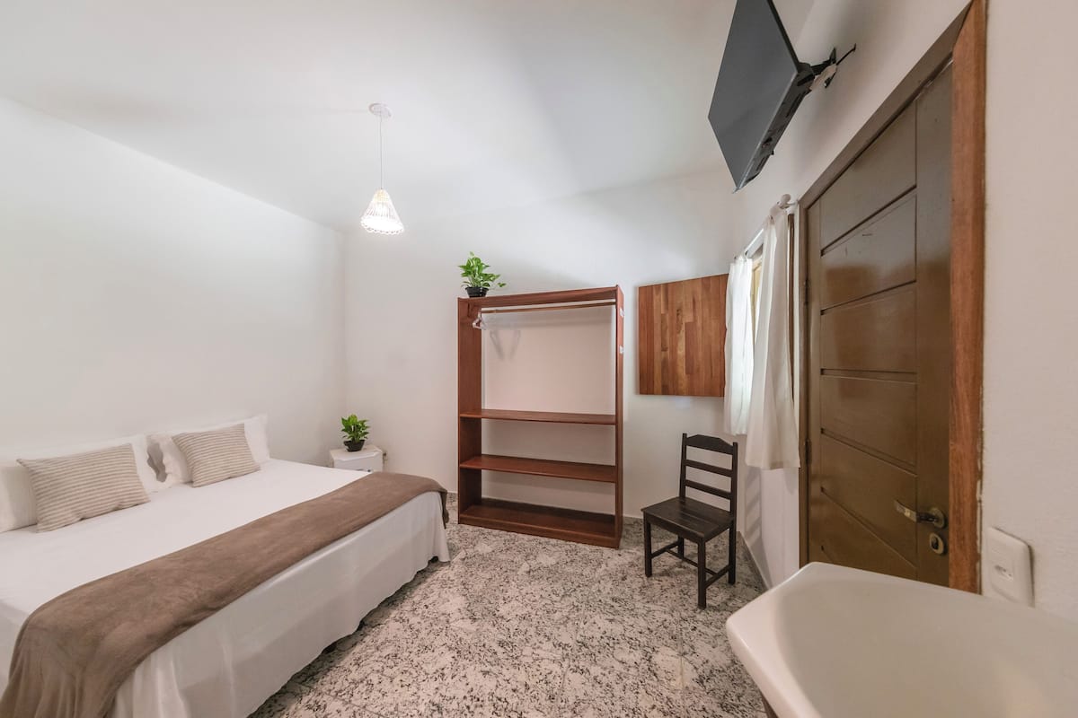 Een slaapkamer met een bed, nachtkastje, hanglamp, houten garderobe, stoel en een bad.