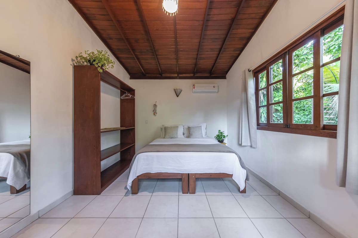 Een slaapkamer met een houten bedframe, witte beddengoed, een houten kledingkast en een raam met uitzicht op groen.