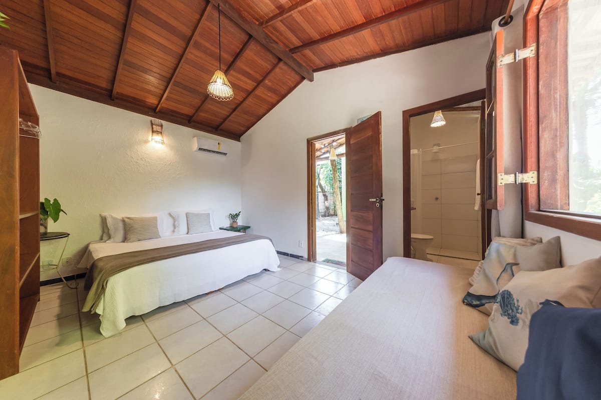 Slaapkamer met houten plafond, een bed met witte beddengoed, nachtkastje met een plant en een deur naar een andere kamer.