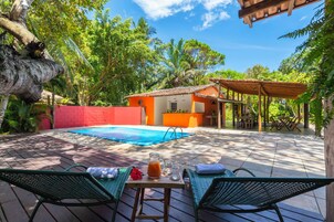 A pool area with lounge chairs, a table with a drink, and a small building with a red wall and a tiled roof.