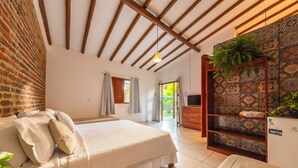 Un dormitorio con cama, techo de madera, pared de ladrillo, piso de baldosas, televisor y una planta.