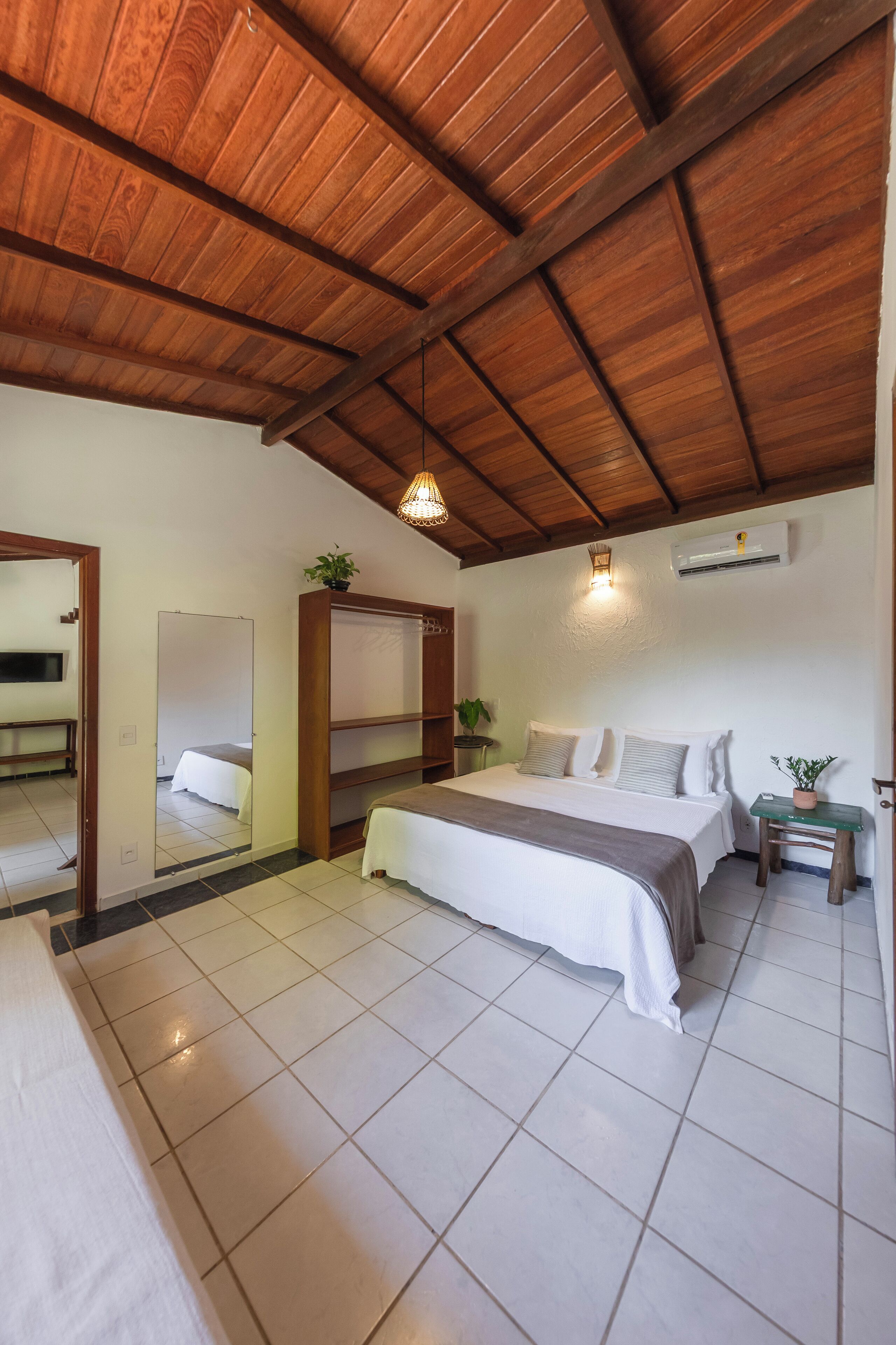A bedroom with a wooden ceiling, a bed with white bedding, a bedside table, a mirror, and a potted plant.
