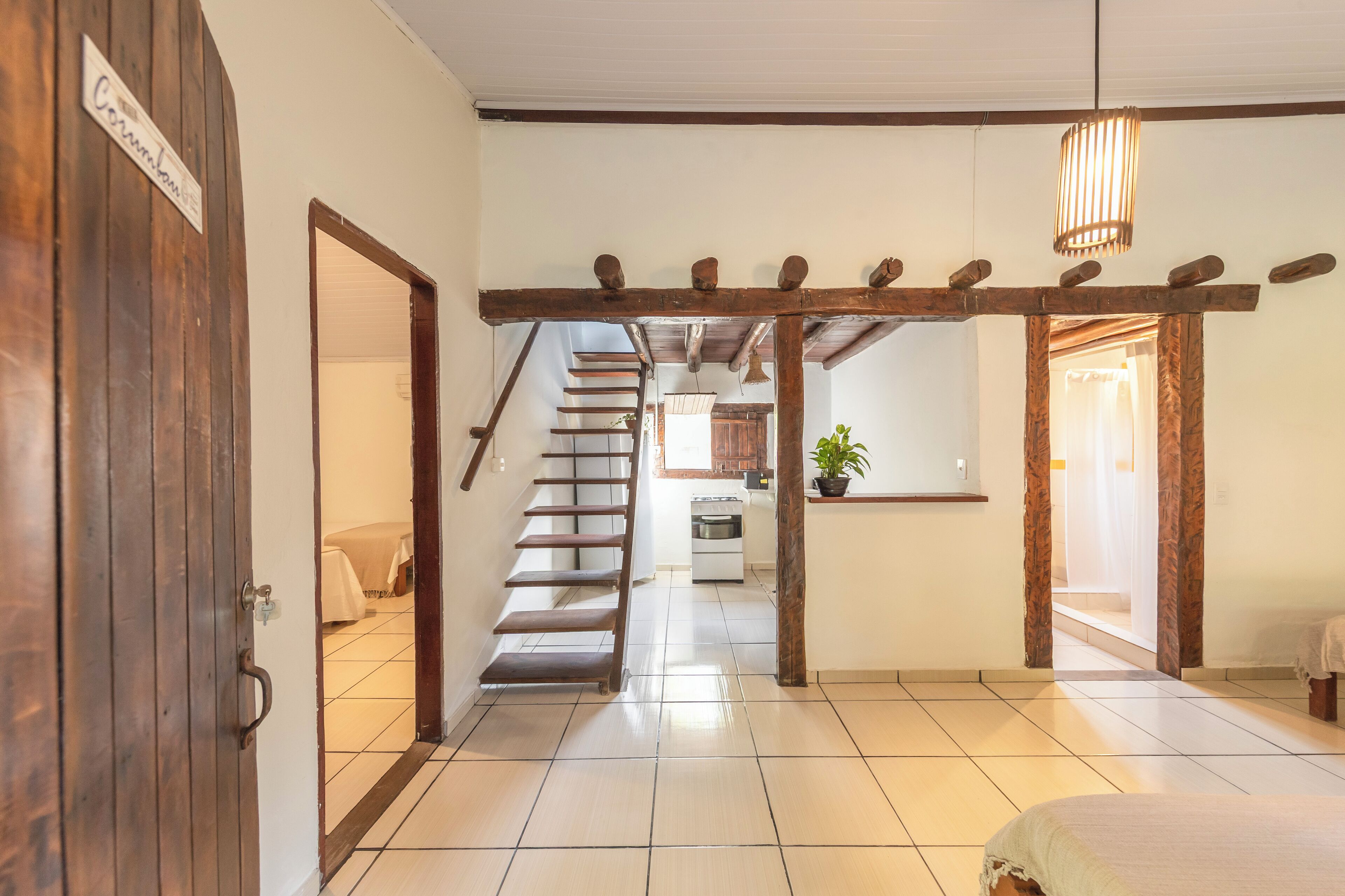 A hallway with a wooden door labeled "Combinado," a staircase, a kitchen area, and a bed with a white bedspread.