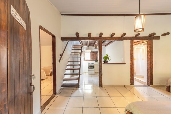 A hallway with a wooden door labeled "Combinado," a staircase, a kitchen area, and a bed with a white bedspread.
