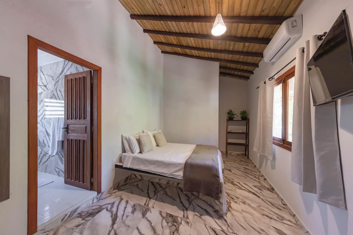 Een slaapkamer met een bed, houten balken in het plafond, een airconditioning, een televisie en een raam met gordijnen.