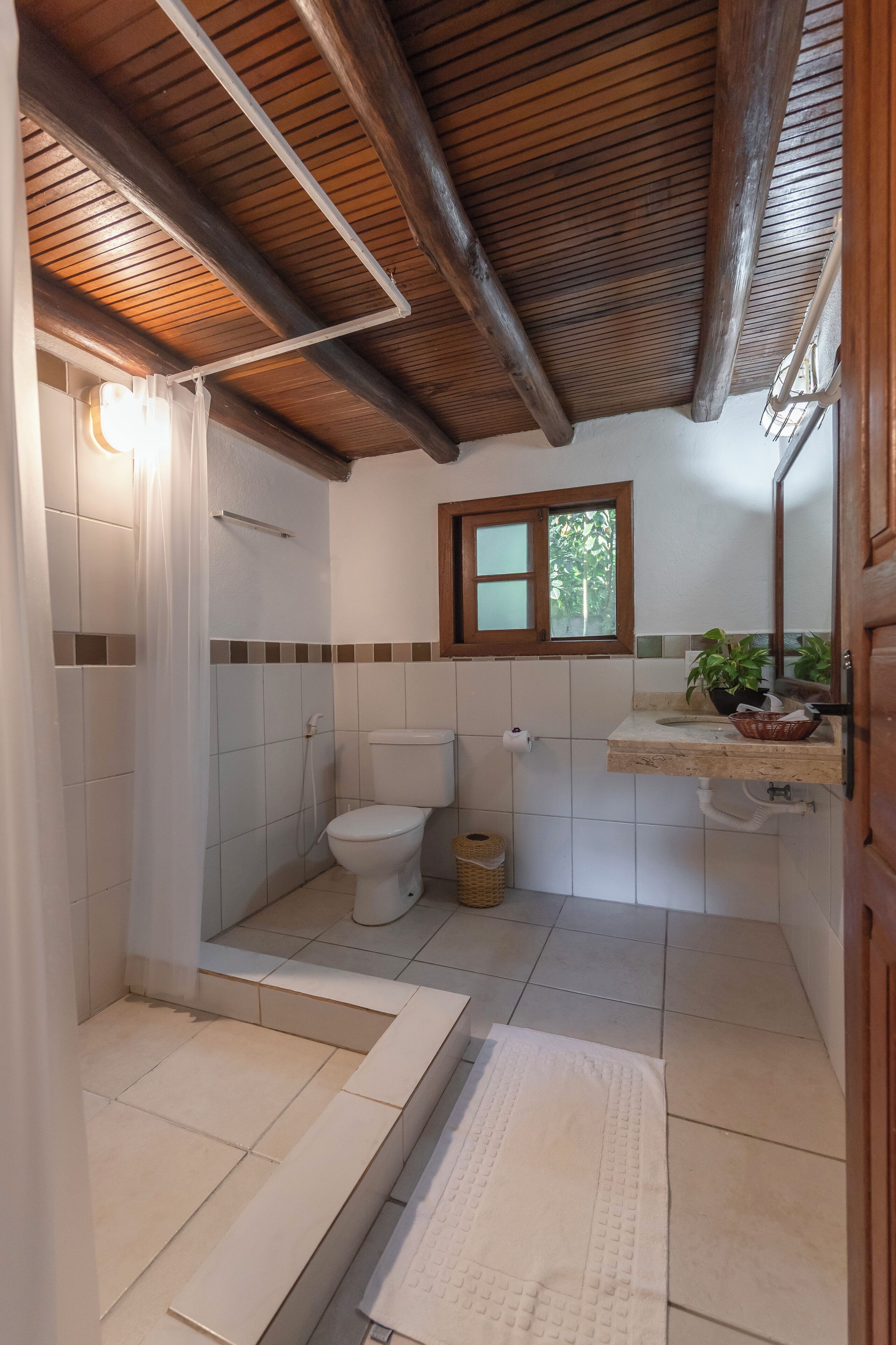 A bathroom with a wooden ceiling, a toilet, a sink with a mirror, and a window.