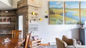 A rustic dining area with a wooden table and chairs, a wooden wall with a door, and a large landscape painting on the wall. - Spotted Fawn Villa | Prime Location in Big Sky Town Center (Big Sky MT)