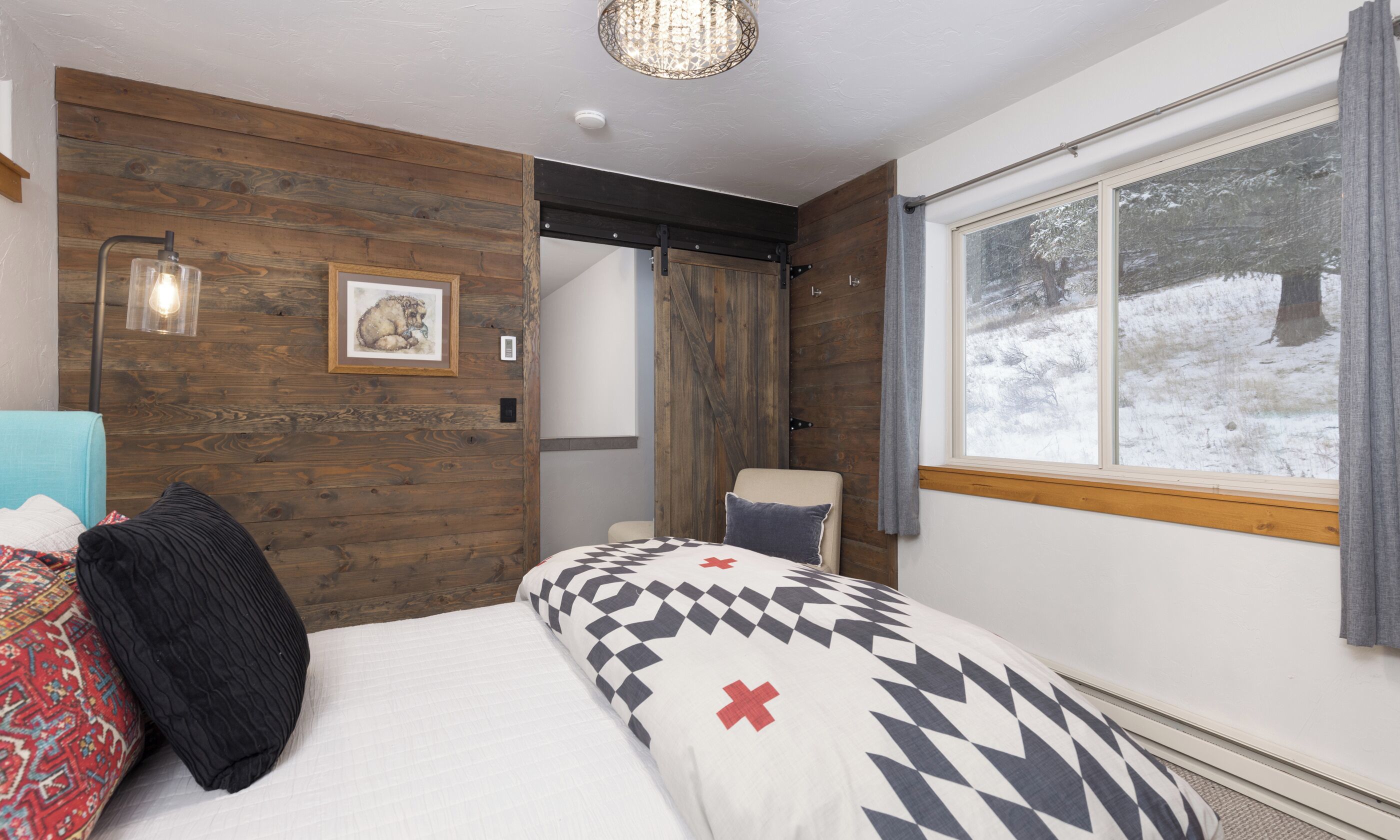 A bedroom with a wooden wall, a bed with patterned bedding, a chair, a window with a view of a snowy landscape, and a hanging light fixture.
