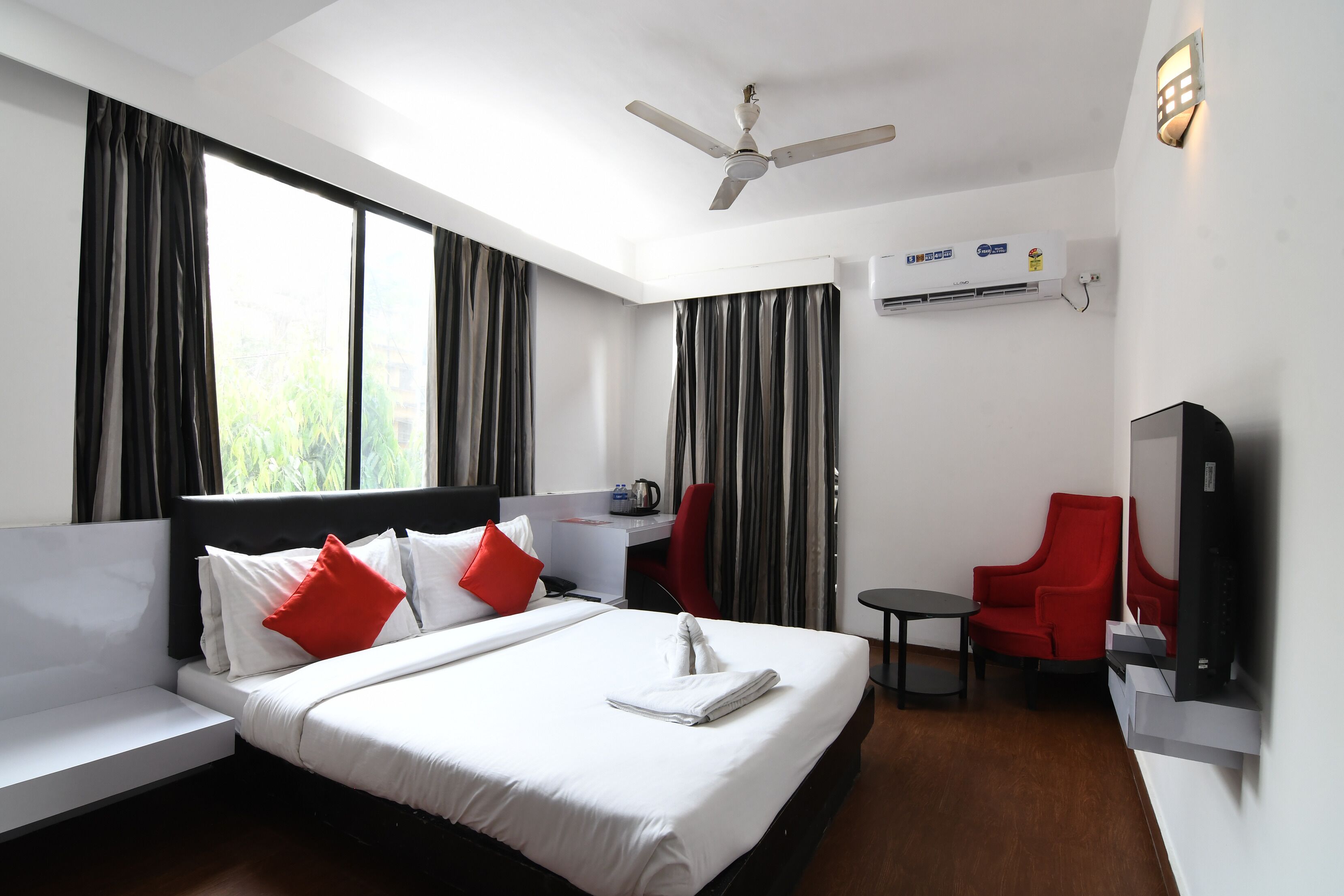 A hotel room with a bed, a desk, a chair, a TV, and an air conditioning unit.