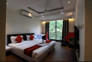 A hotel room with a large bed, a desk, and a chair. There is a window with curtains and a view of greenery outside.