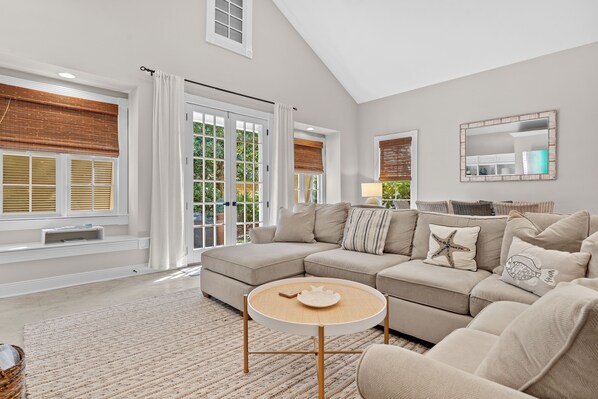 A spacious living room with a large sectional sofa, a round coffee table, and a view of the outdoors through large windows. - Sea La Vie 3 Bedroom Home (Miramar Beach)