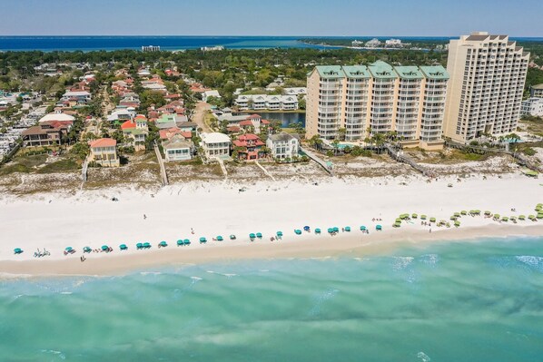 An aerial view of a coastal area with a beach, a row of beach umbrellas, and several multi-story buildings. - Sea La Vie 3 Bedroom Home (Miramar Beach)