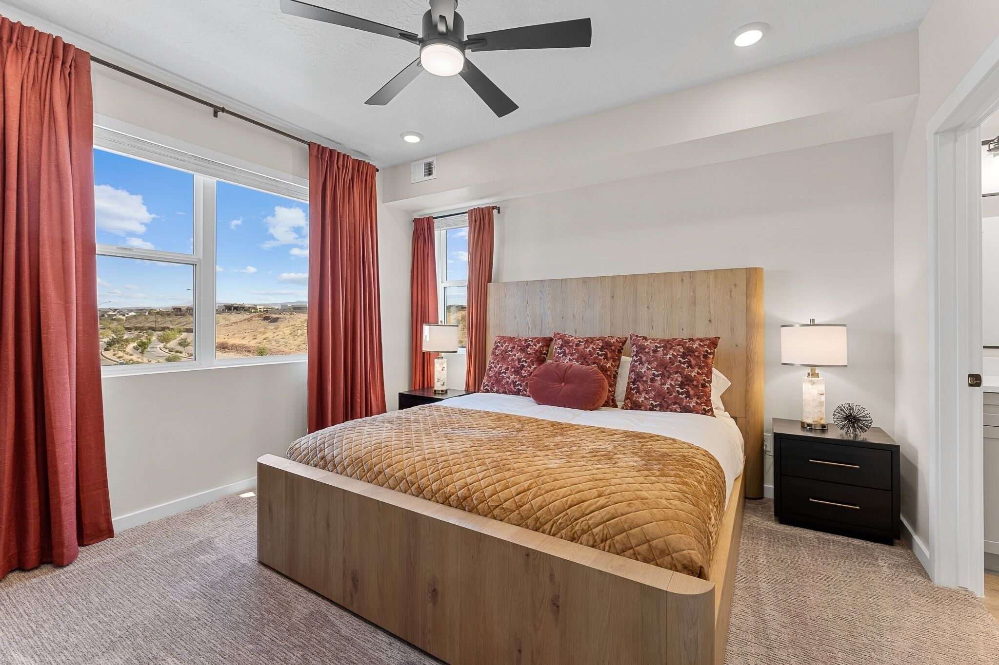 A bedroom with a large bed, a ceiling fan, a nightstand, and a view of the desert landscape.