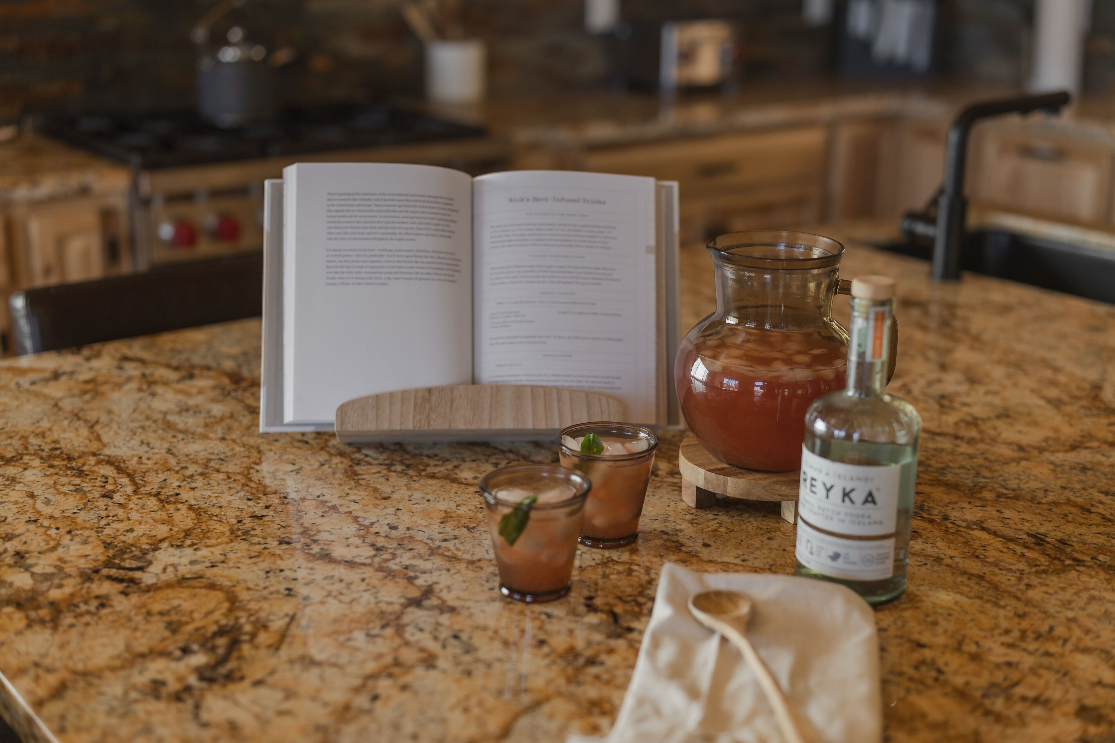 A kitchen counter with a book, two glasses of iced tea, a bottle of liquor, and a small bag.